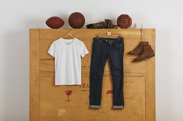 Белая пустая хлопковая рубашка представлена рядом с джинсами japanese selvedge и кожаной обувью на деревянном большом грузовом ящике с игровыми мячами vitage наверху