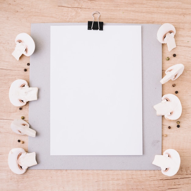 Бесплатное фото Белый чистый лист бумаги прикреплен в буфер обмена, украшенный половинками грибов и черного перца на деревянном текстурированном фоне