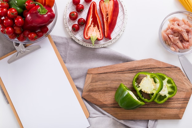 White blank paper on clipboard with bell pepper; cherry tomatoes and chicken on white backdrop