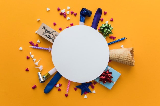 White blank round frame over the party blower; gift box; candles; sprinkles and waffle cone against an orange colored backdrop