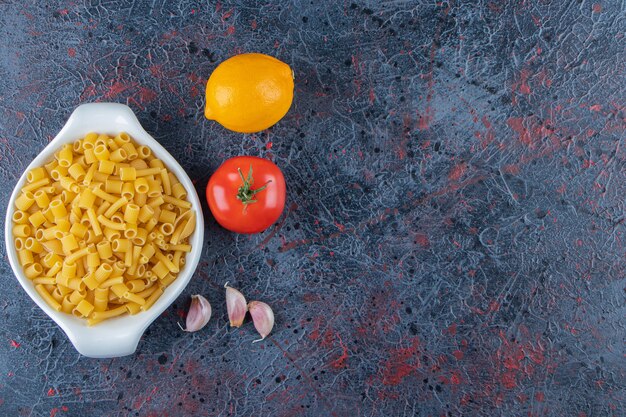 A white board of raw pasta with fresh red tomatoes and lemon on a dark background . 