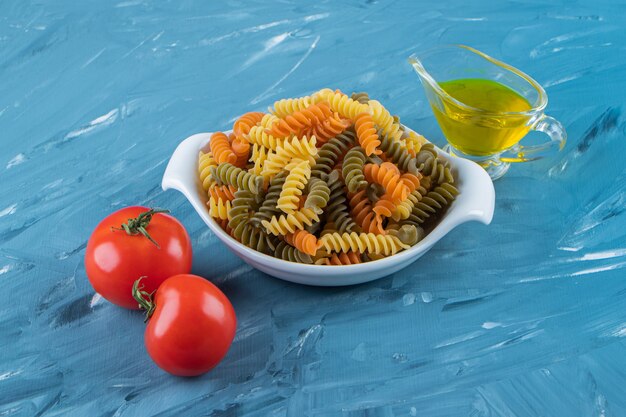 A white board of raw pasta with oil and fresh red tomatoes on a blue surface.