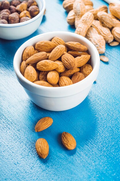 White bowl of almonds; peanuts and peanuts on blue textured backdrop