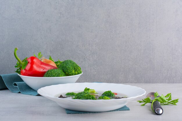 White bowl of fresh vegetable soup and organic vegetables on stone surface