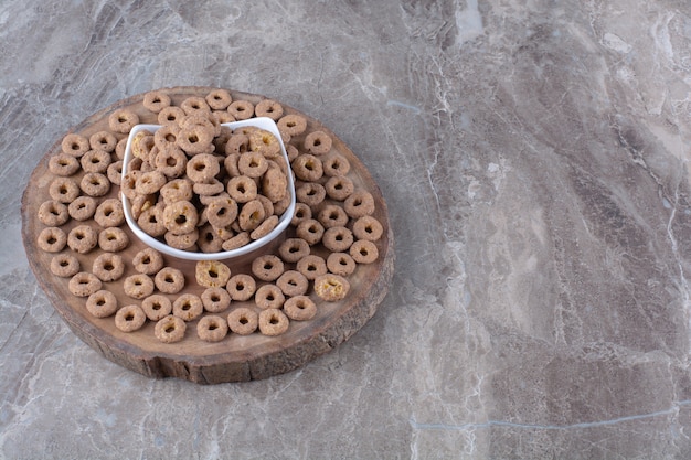 A white bowl full of healthy chocolate cereal rings on a wooden piece . 