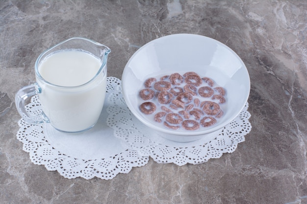 Una ciotola bianca di anelli di cereali al cioccolato sani con una brocca di vetro di latte.