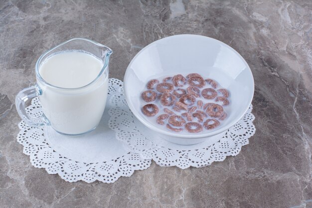 A white bowl of healthy chocolate cereal rings with a glass jug of milk.