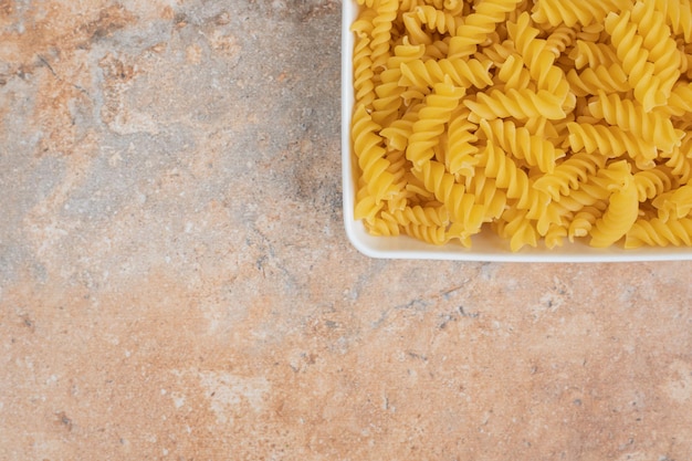 A white bowl of unprepared macaroni on marble space. 