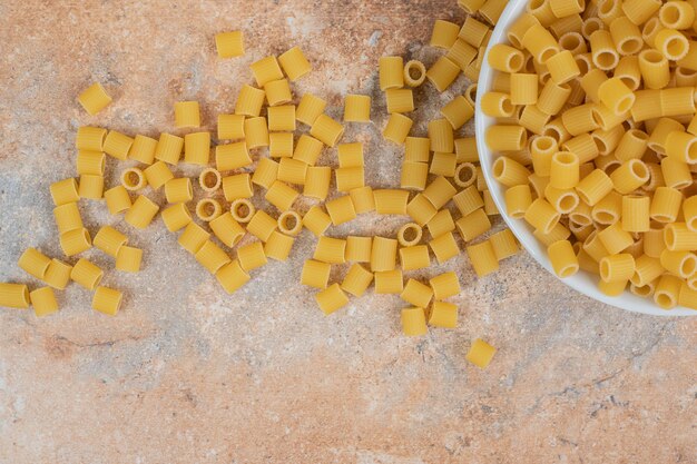 A white bowl of unprepared macaroni on marble space. 