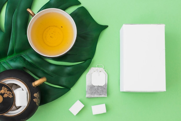 White box, herbal tea, sugar cubes and tea bag on green paper background