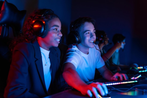 Free photo white boy and black girl teens in headsets playing video games in video game club with blue and red illumination keyboard and mouse with illumination