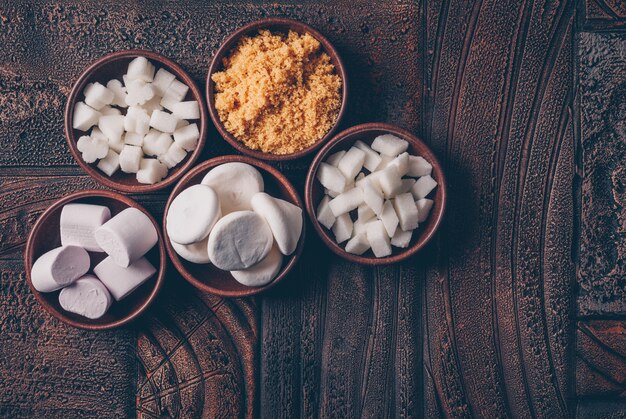 White and brown sugar in bowls with candies and marshmallow top view on a dark wooden table