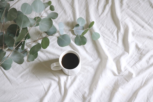 White Ceramic Mug with Coffee Next to Silver Dollar Gum Tree Leaves on White Bed-sheet