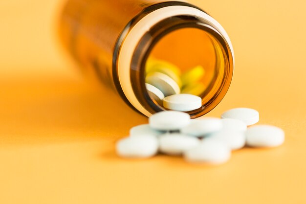 White circular pills spilling from glass bottle over the yellow background