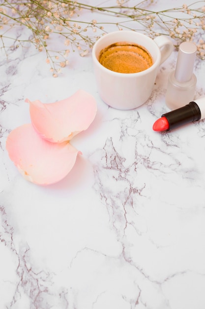 White coffee cup; rose petals; nail polish bottle; baby's-breath flowers and lipstick on white textured backdrop