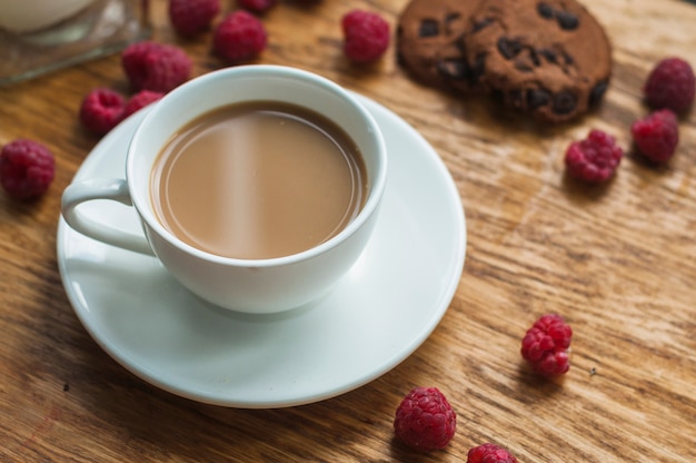 木製の背景にチョコレートクッキーとラズベリーとコーヒーの白いカップ