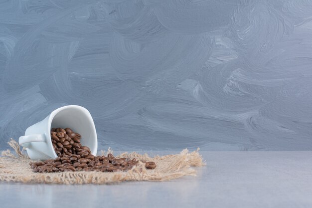 A white cup of roasted coffee beans on marble.