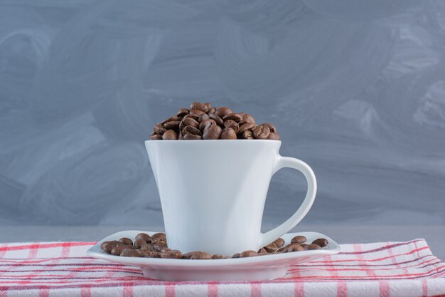 A white cup of roasted coffee beans on striped tablecloth.