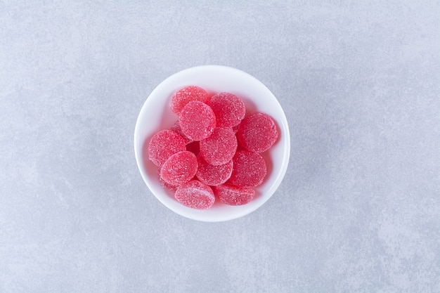 A white deep plate full of red sugary fruit jelly candies on gray surface