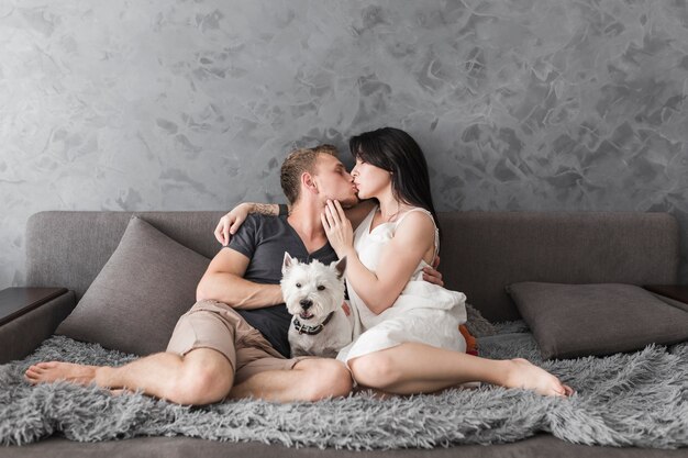 Белая собака между молодой парой, сидящей на диване поцелуи