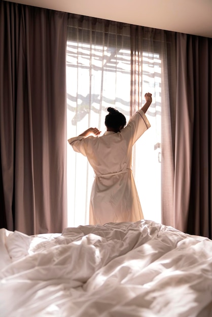白いドレスアジアの女性の美しい女性のストレッチ朝は、白いカーテンと太陽の輝きの背景のライフスタイルの家のコンセプトと居心地の良いインテリアの寝室で新鮮さとのんきな週末の瞬間を目覚めます