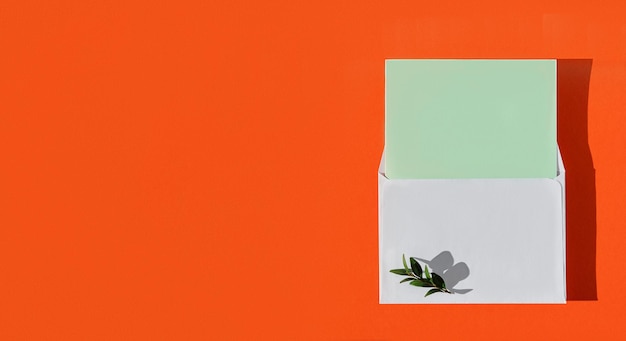 Free photo white envelope with green blank card and green leaves on burnt orange background, top view with copy space. minimalistic mockup with envelope and blank card for congratulations, flat lay