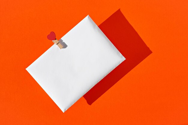 White envelope with a red heart on a clothespin, on an orange background. Love letter or Congratulations on Valentine's Day, Mother's Day, Women's Day. View from above
