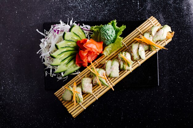 White fish fillet on sushi mat with ginger, wasabi and cucumber on black table.