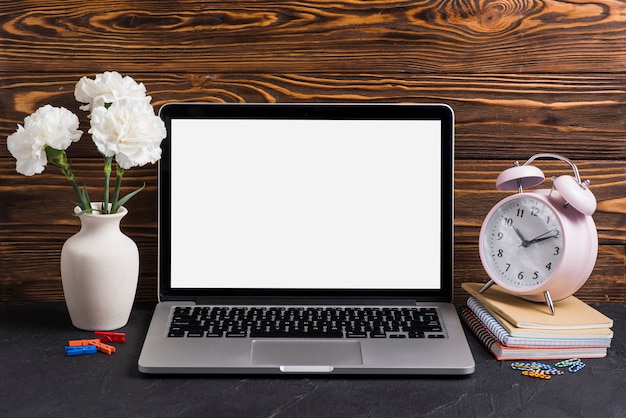 White flowers in the vase; laptop and alarm clock on notebooks against wooden backdrop