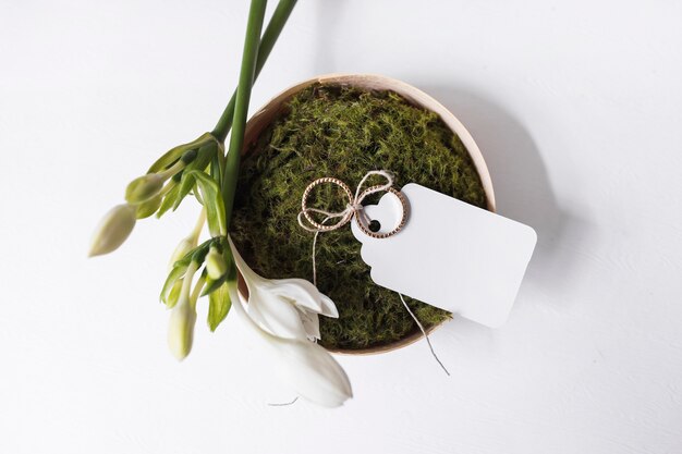 White flowers and wedding rings with blank tag on moss bowl over white background