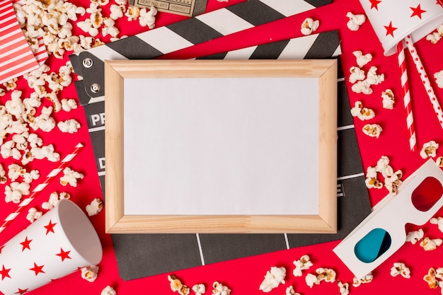 Free photo white frame over the clapperboard with popcorns; drinking straws and 3d glasses on red backdrop