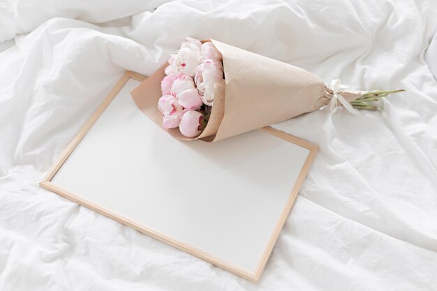 White frame mockup on the bed Bouquet of pink peonies in craft packaging Scandivanavian white interior