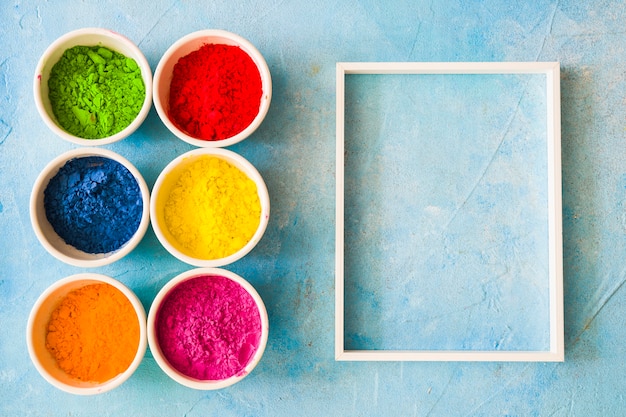 Free photo white frame with holi color powder in the bowl on painted blue backdrop