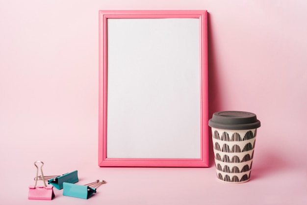 Free photo white frame with pink border; paper clips and coffee disposable cup against pink background