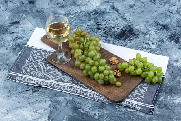White grapes, walnuts on a cutting board with kitchen towel, glass of whisky high angle view on a dark blue marble background