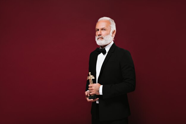 White haired man in suit looks into camera and holds Oscar statuette