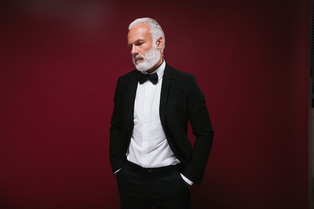 White haired man in suit poses on burgundy wall