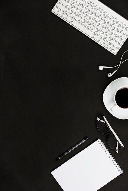 White keyboard; earphone; coffee cup; eyeglasses; pen and spiral notepad against black desk