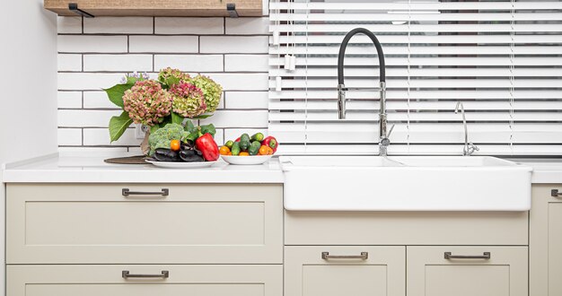 White kitchen cabinets with metal handles on the doors near the washbasin with a bouquet of flowers and a plate of vegetables.