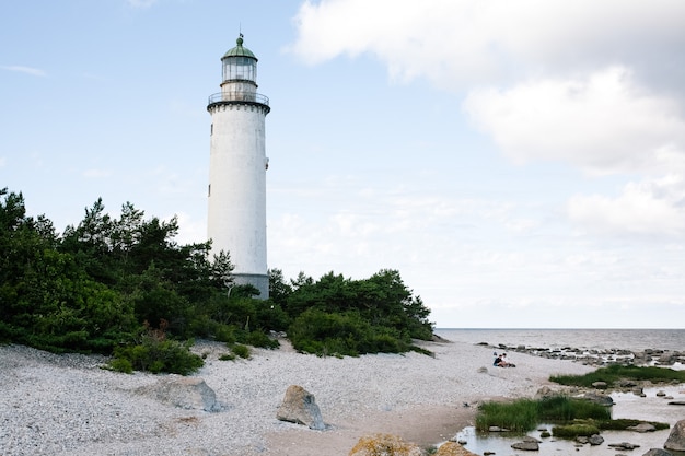 Белый маяк в окружении деревьев у берега пляжа с облачным небом