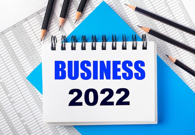 White notebook with the text business 2022 on the table next to black pencils on a blue background and reports. business concept