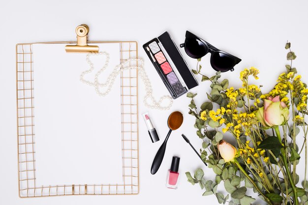 White paper on clipboard; necklace; sunglasses; lipstick; nail varnish bottle; makeup brush and flower bouquet on white background