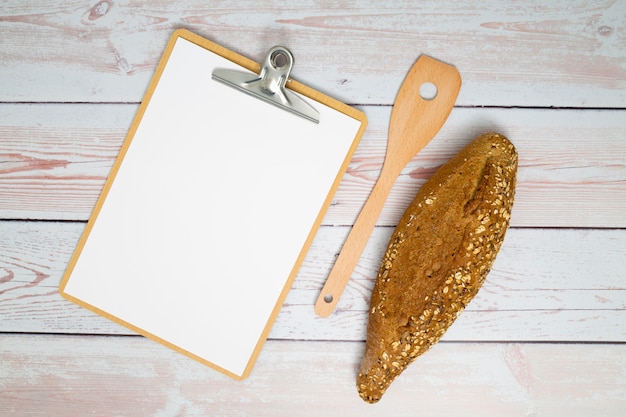 White paper on clipboard; spatula; and loaf of bread on wooden desk