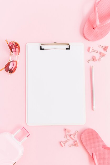 White paper on clipboard surrounded with miniature luggage bag; sunglasses; paperclips; pencil and flip flops on pink background