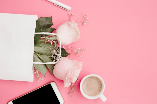 Foto gratuita penna bianca; rose nella borsa della spesa; smartphone e tazza di caffè su sfondo rosa