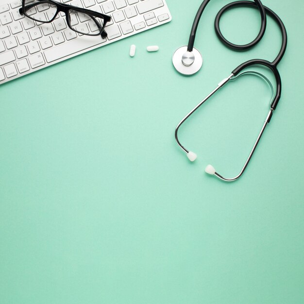 White pills and stethoscope near spectacles on wireless keyboard over backdrop