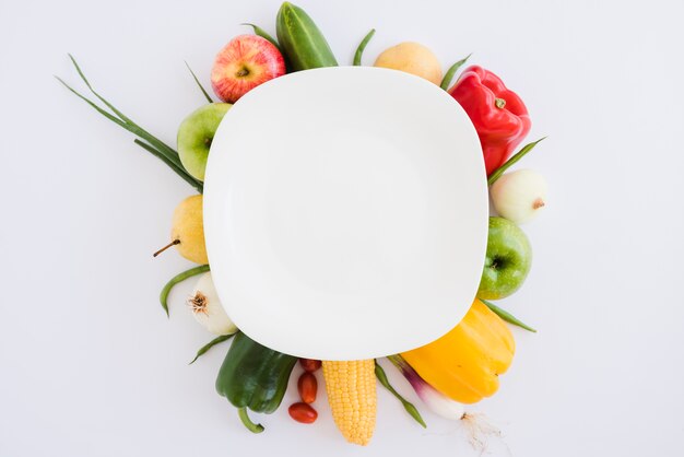 きゅうりの上の白い皿。林檎;ピーマン;玉ねぎ;トウモロコシと白い背景の上のネギ