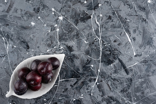 White plate of fresh purple plums on marble surface.