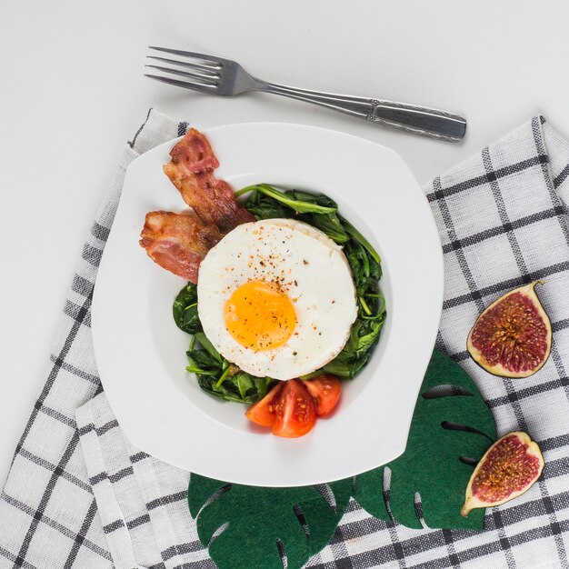White plate of fried eggs; spinach; tomatoes and bacon over the white background with napkin and fork
