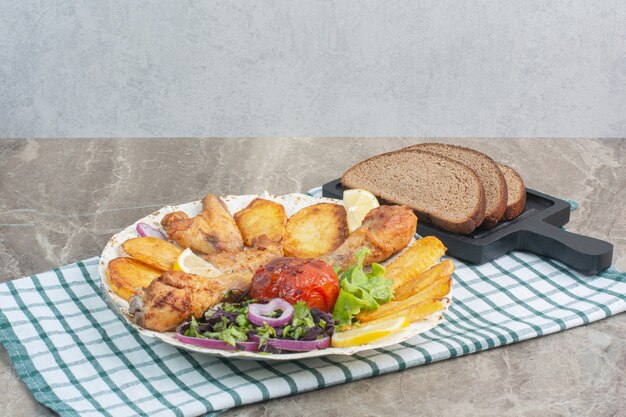 Белая тарелка, полная жареного цыпленка и картофеля с черным хлебом.